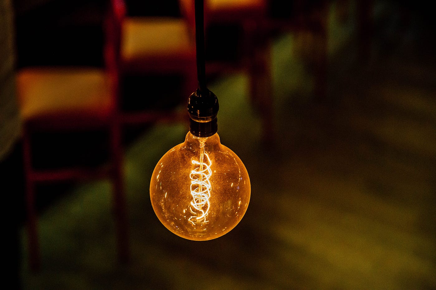A close-up of a lit vintage-style lightbulb hanging from a black cord, revealing a bright, glowing spiral filament inside. The background is dark and out of focus, showing faint warm lights and silhouettes of chairs.