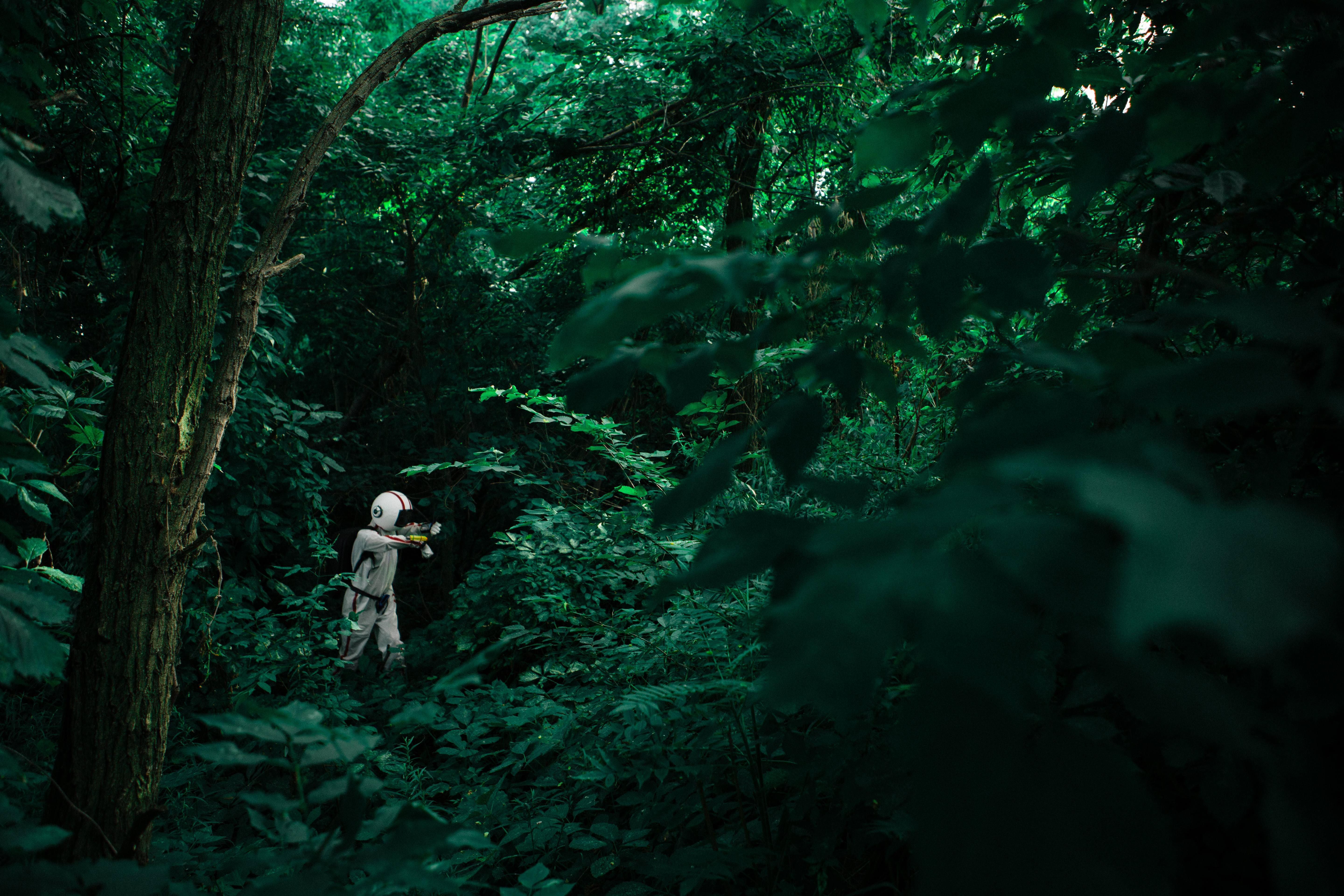 A person wearing a full white spacesuit and helmet stands partially obscured within a dense, lush green forest. The deep green foliage surrounds the figure, creating a stark contrast with the bright white suit.