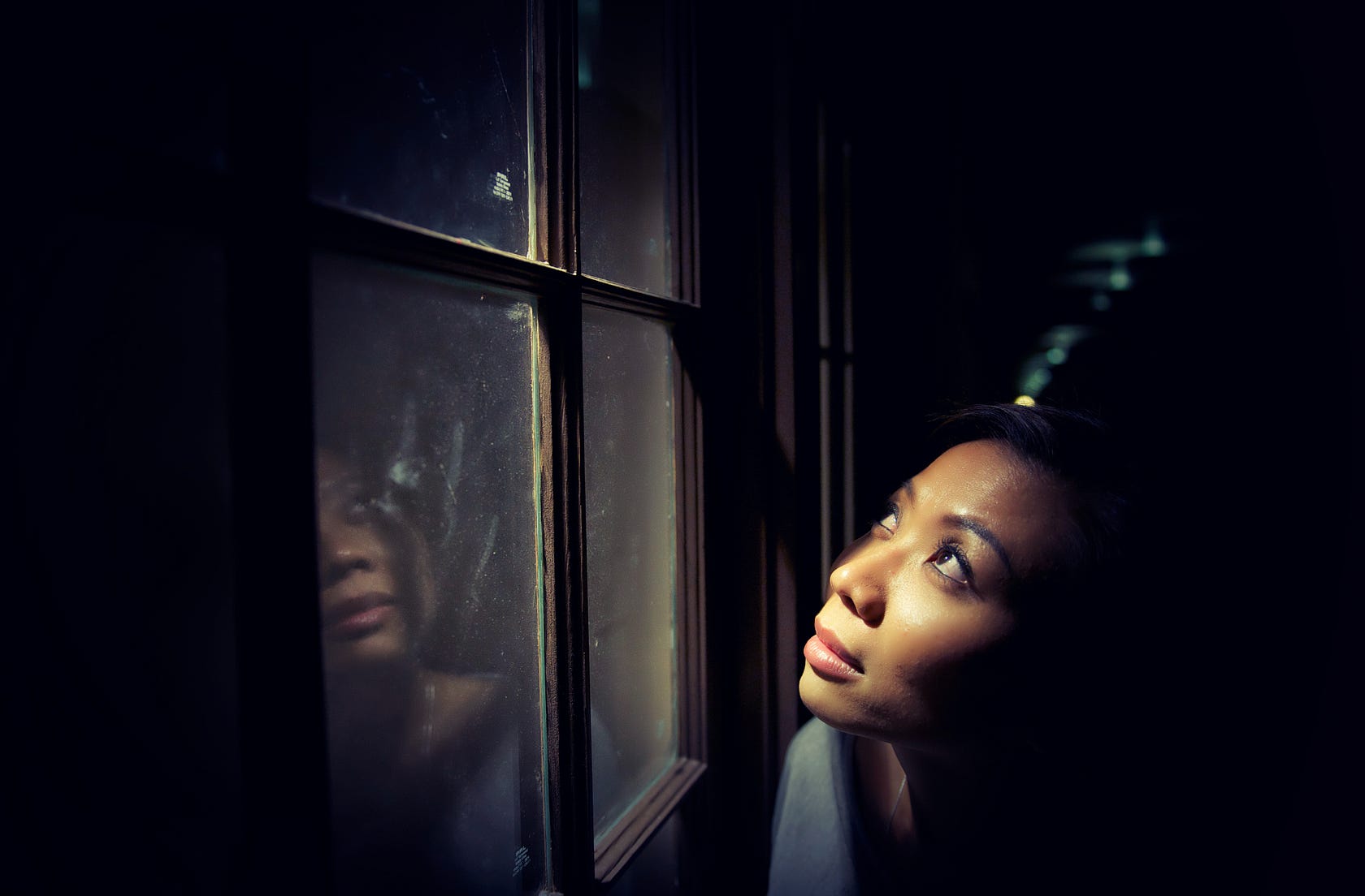 A woman stands in the dark looking upward next to a window, her face clearly illuminated by a warm light source. Her faint reflection is cast onto the dark window glass beside her.