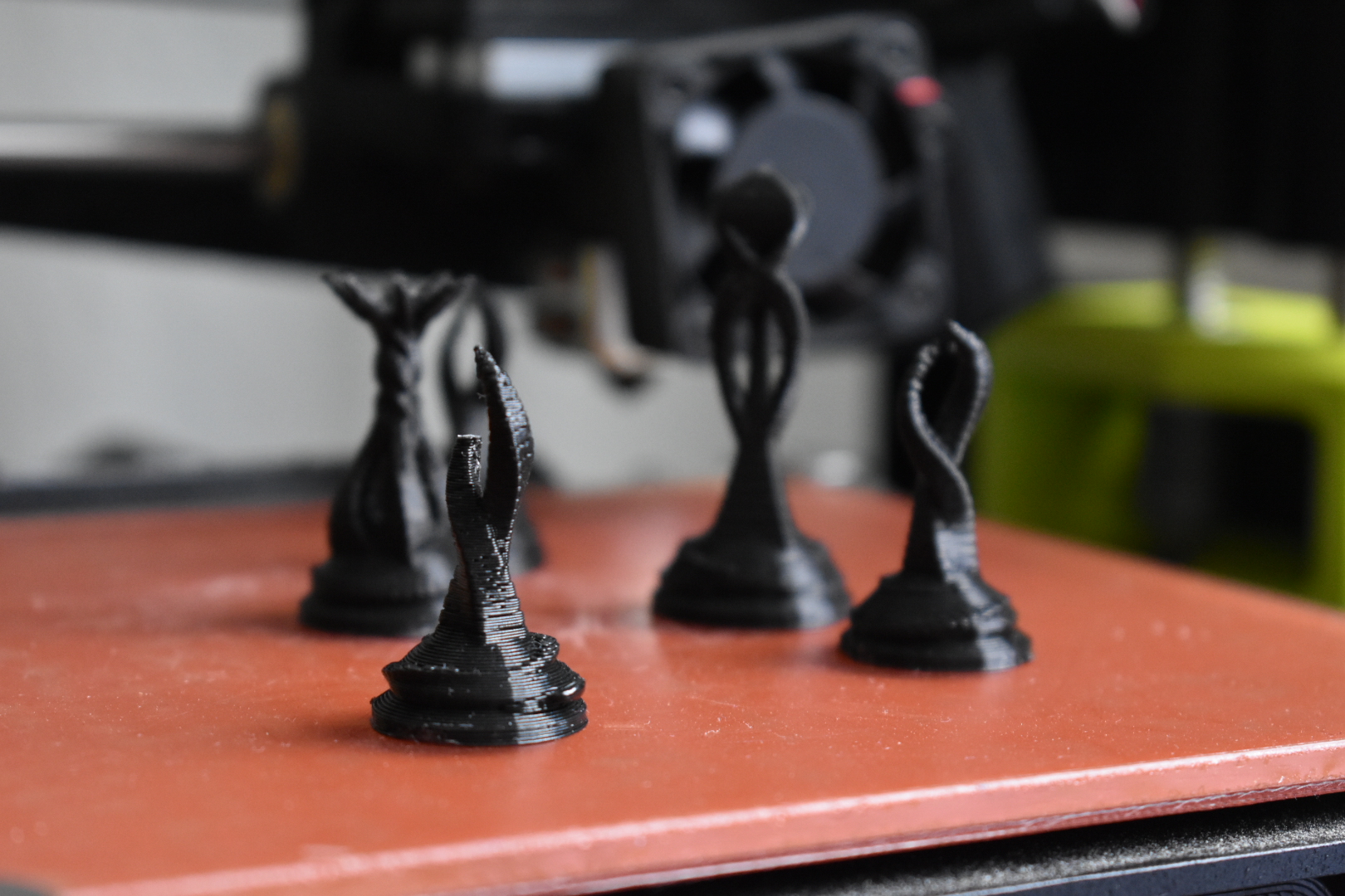 Four chess pieces sit atop a square, orange plate. The chess pieces are made with filament from a 3-D printer. Faded out in the background, there is the head of a 3-D printer, presumably what made the chess pieces. This photo was taken at Open Labs Hackerspace in Tiranë, Albania.