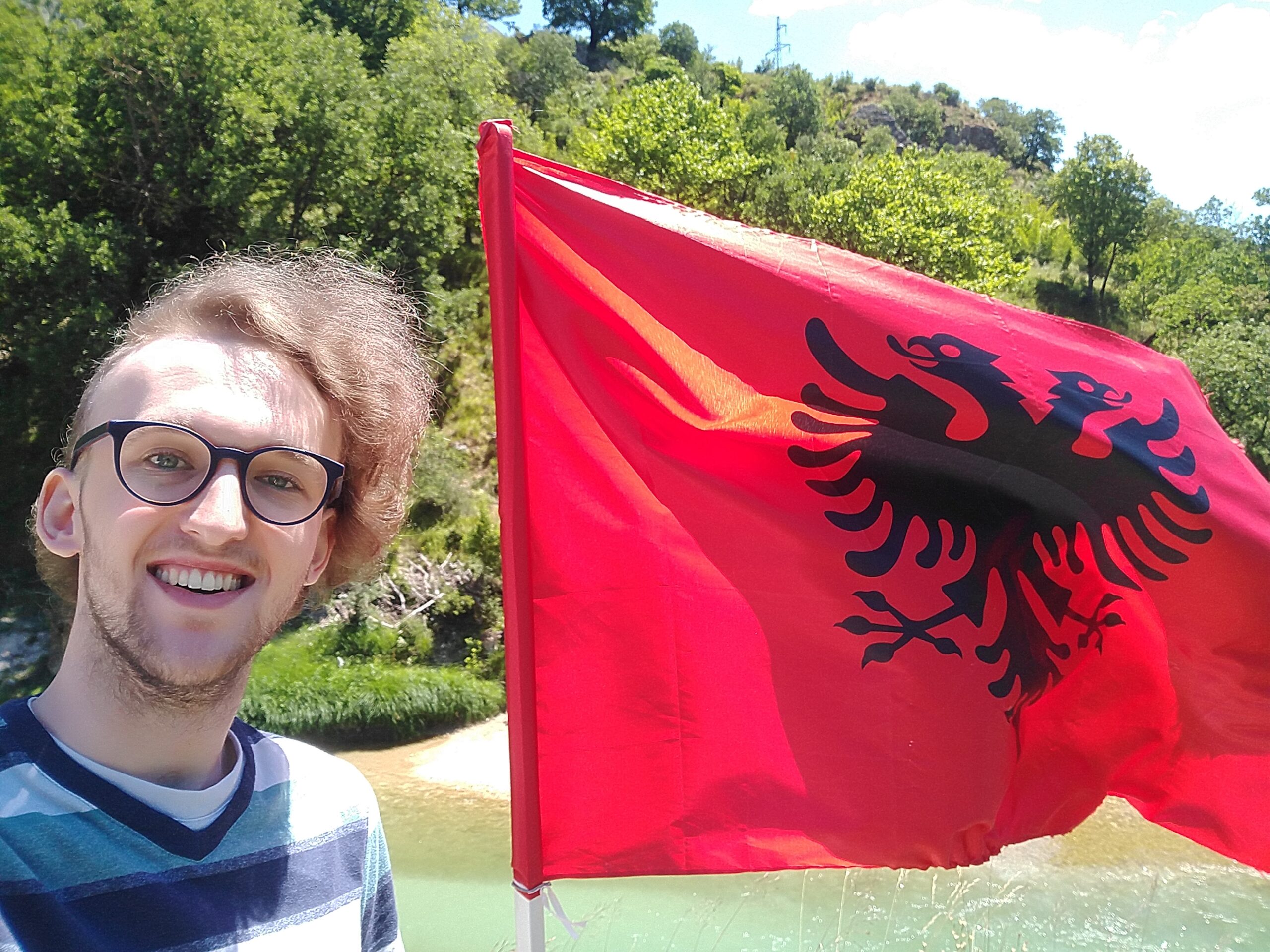 Justin is pictured to the left of the Albanian flag, adjacent to his head. In the background, there is a small river surrounded by trees and small sand beaches.