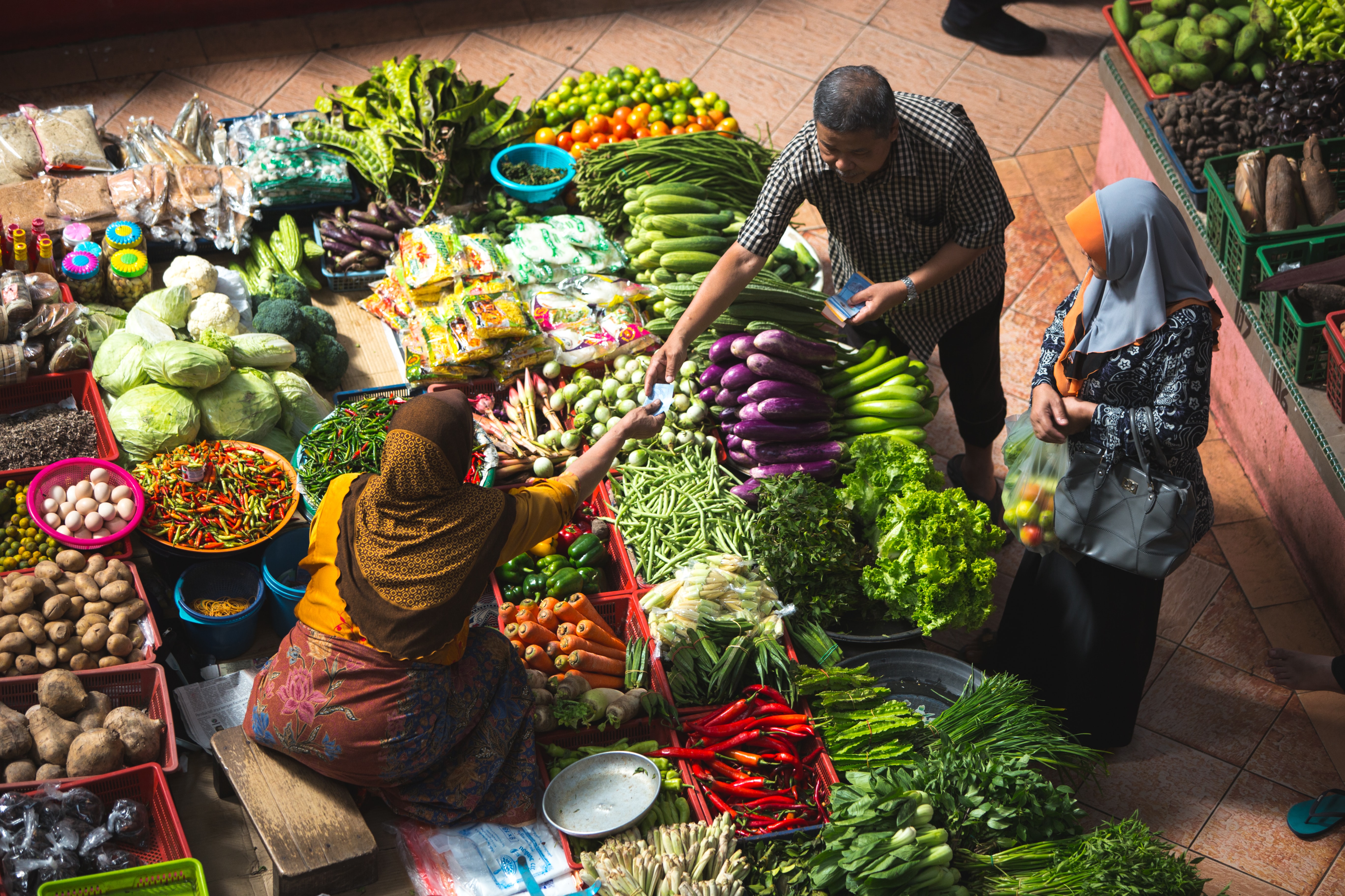 View looking down at a small farmer&rsquo;s market, where a woman sits behind several different cases of vegetables. A man hands payment to the woman for unseen goods. This is connected how knowing your customers can also be like knowing your community.