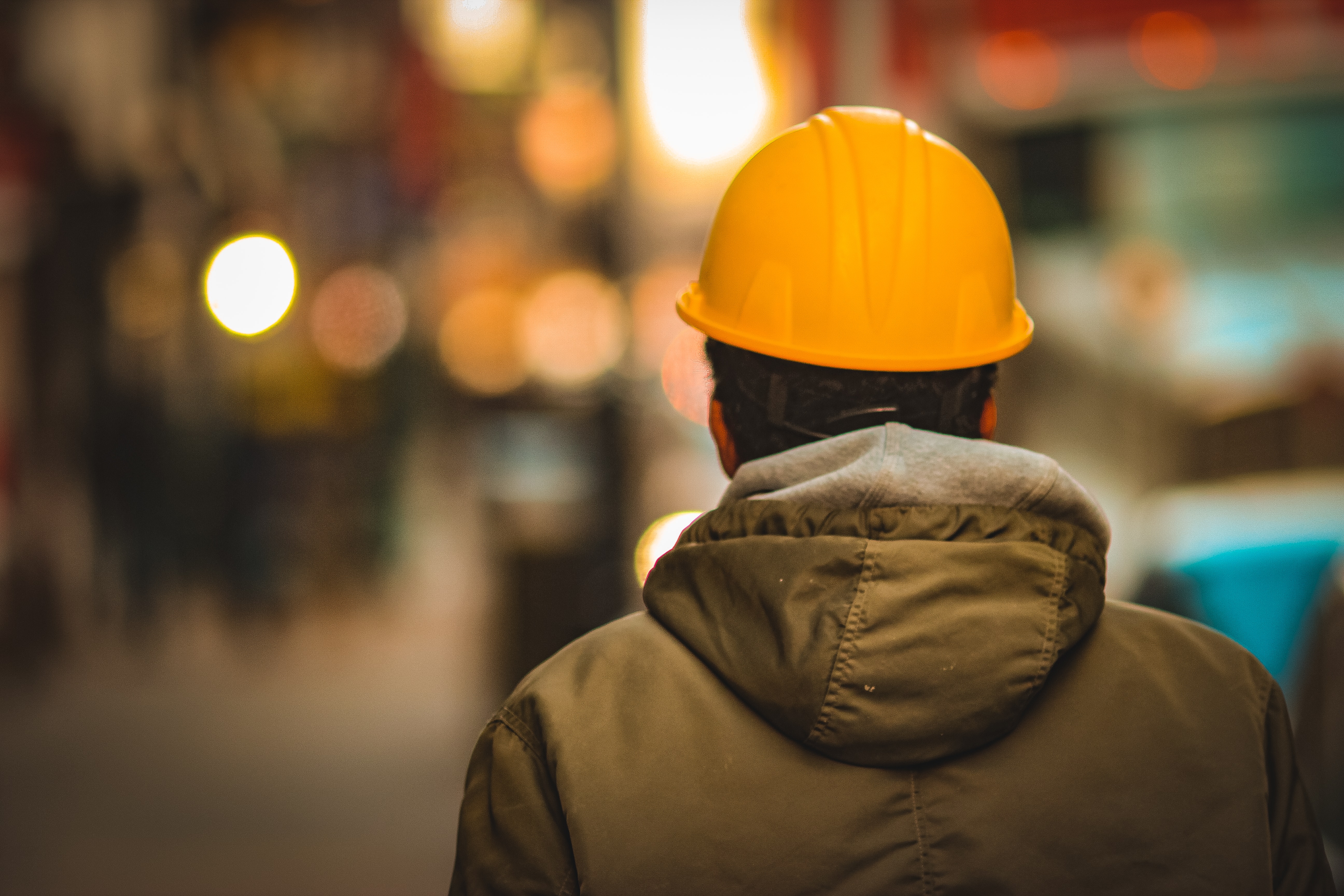 A man is facing forward with his back to the camera. He wears a heavy coat and a construction hard hat. The background is blurred and unclear. In this way, we can think of labor investment from a human-centered approach first.