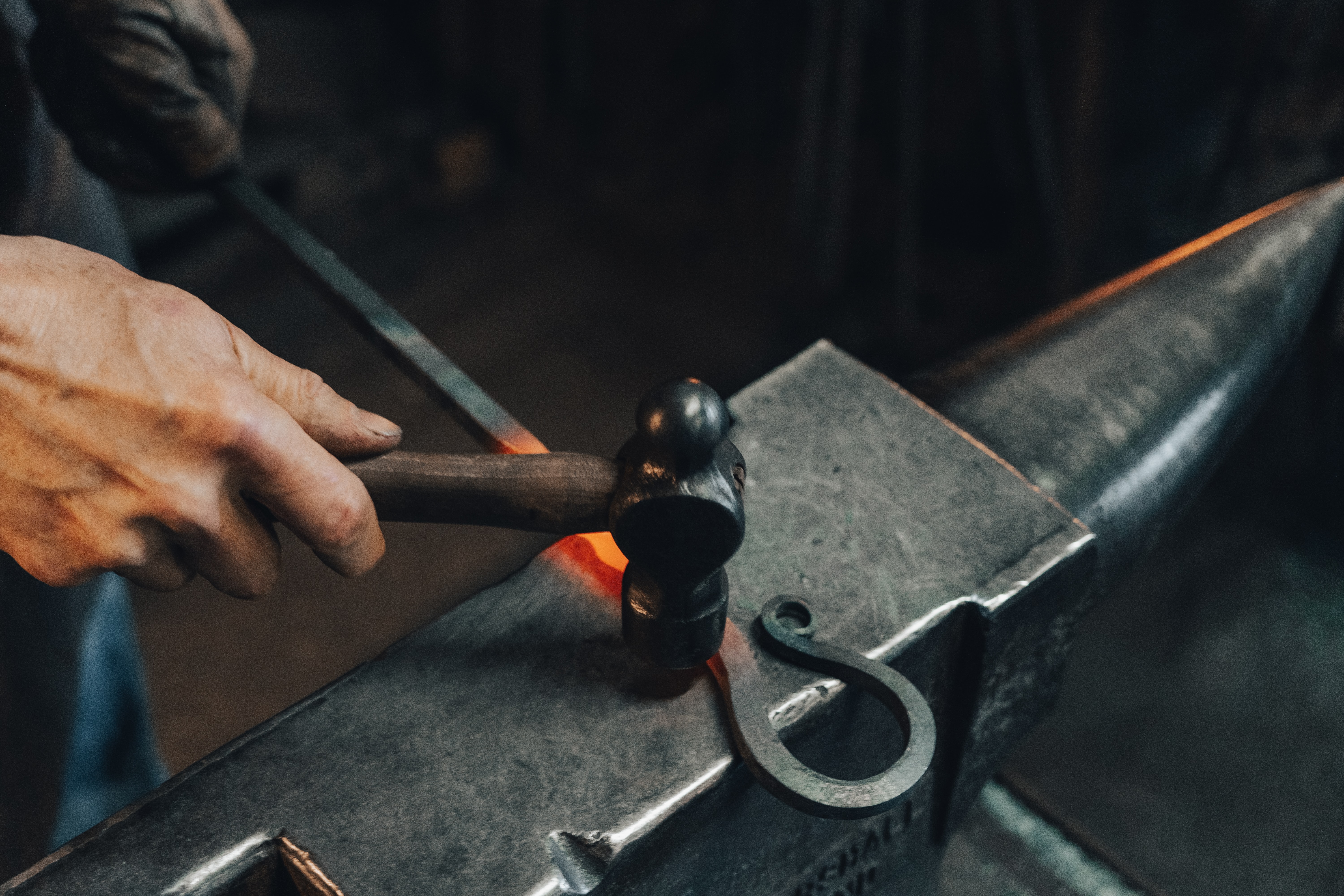 A metalworker is working at an anvil. A red-hot iron rod is on the anvil, and a person uses a hammer to shape and mold the hot iron into a hooked shape.
