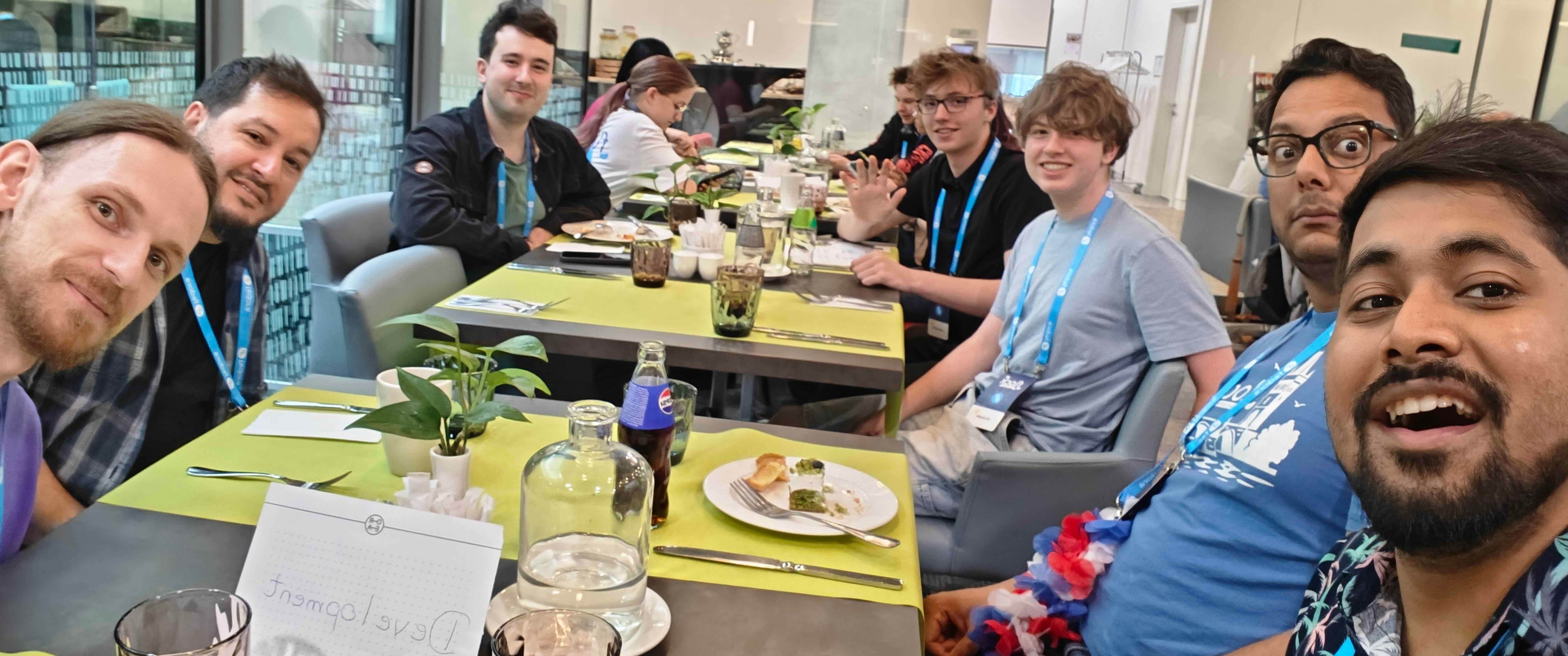 A group selfie of several individuals wearing blue Flock to Fedora conference lanyards gathered around a long dining table in a room with large glass windows. A handwritten sign reading "Development" sits on the table among plates and glasses as the group poses for the camera held by a man in the foreground.