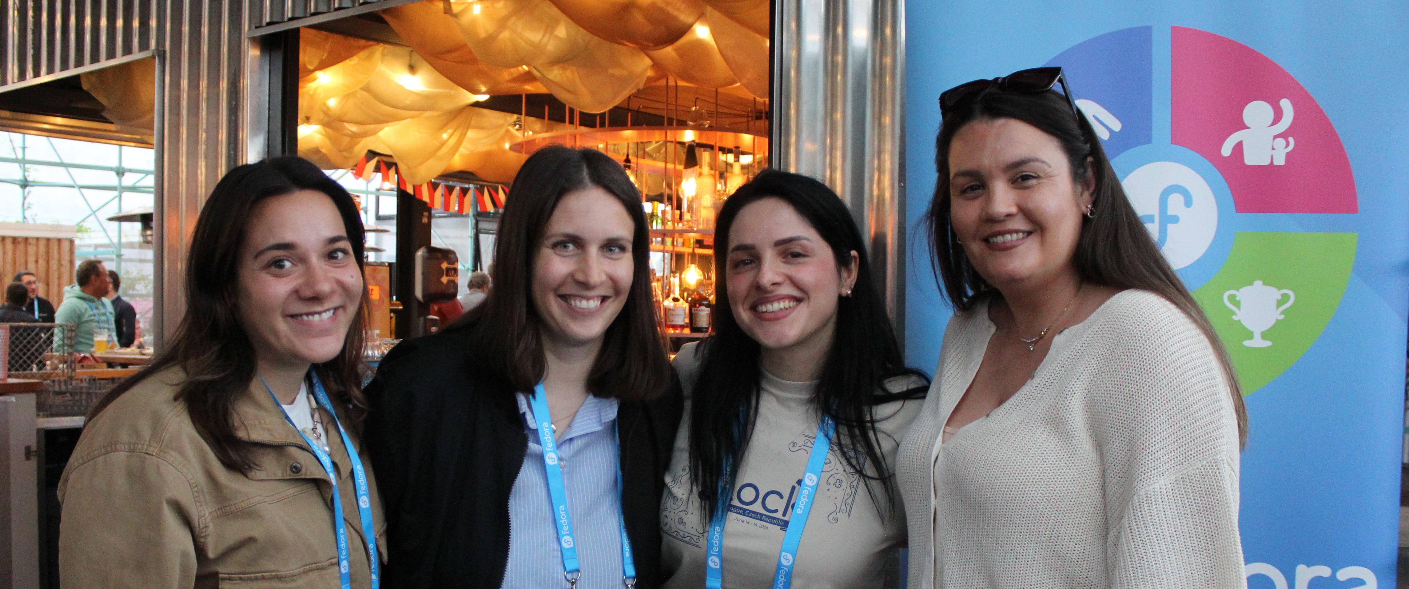 A photograph of four smiling Flock to Fedora organizers posing together at the Monday night social reception. They are standing in front of a warmly lit bar area and a large blue Fedora graphic banner. Left to right: Jennifer Schimmoller, Dorka Volavkova, Jona Azizaj, Aoife Moloney.