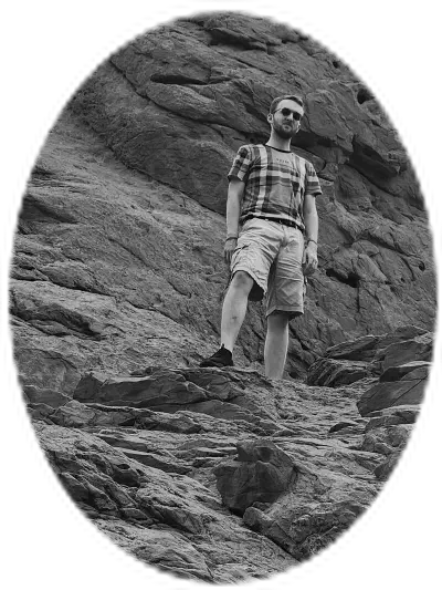 A photo of the Garden of the Gods in Colorado Springs, Colorado, United States. Towering red rock formations stand tall against a blue sky with white clouds. A man stands higher up in the rocks, wearing cargo shorts and a plaid t-shirt.